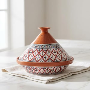 May include: A terracotta tagine featuring a conical lid and a wide base. The exterior is decorated with a hand-painted geometric pattern in red, black, blue, and green colors over a white background, with a small steam vent at the top.