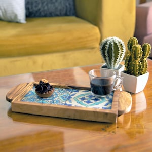 Op de afbeelding: Een houten dienblad met een blauw en wit betegeld ontwerp. Het dienblad staat op een houten tafel met een kopje koffie en een klein dessert erop. Er staan twee kleine cactussen in witte potten op de tafel.