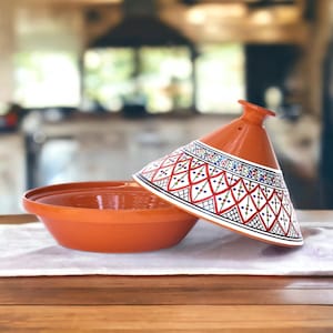 May include: A terracotta tagine with a white lid featuring a geometric pattern in red, blue, and green. The tagine is a traditional Moroccan cooking pot.