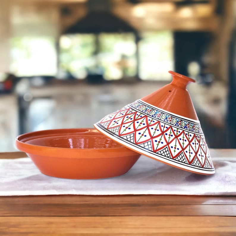 May include: A terracotta tagine with a white lid featuring a geometric pattern in red, blue, and green. The tagine is a traditional Moroccan cooking pot.