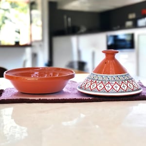May include: A terracotta tagine with a white and blue geometric pattern. The tagine has a round base and a conical lid. The lid has a small hole in the top.