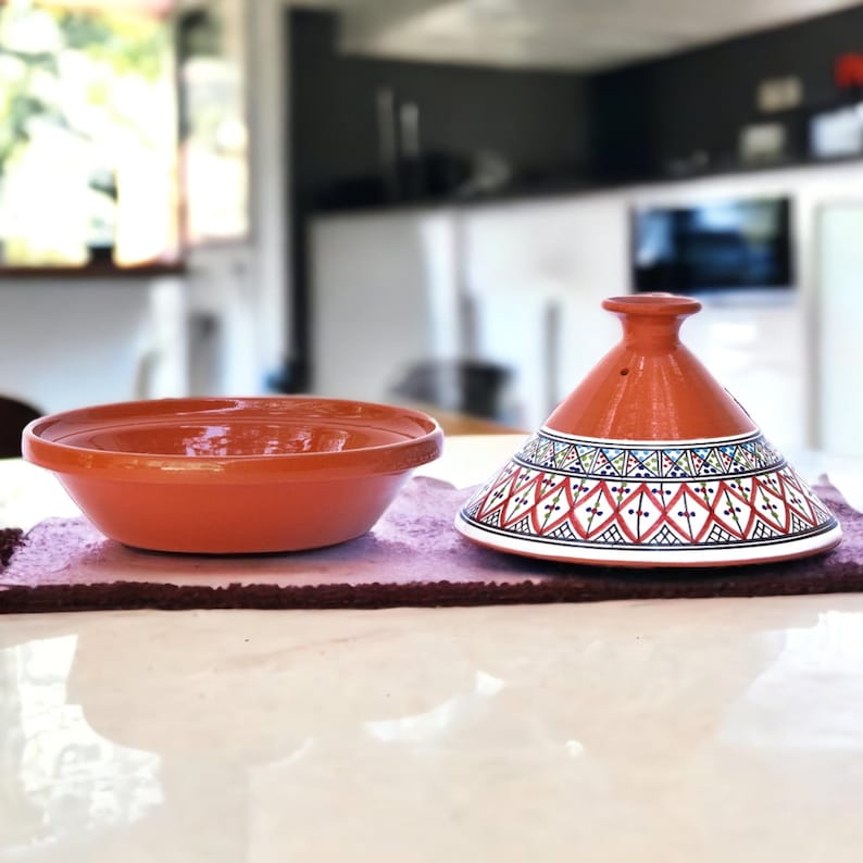 May include: A terracotta tagine with a white and blue geometric pattern. The tagine has a round base and a conical lid. The lid has a small hole in the top.