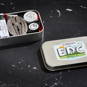 May include: A silver metal tin with a black and white sticker that says "KOUKOU SKYETA EDC -SURVIVAL KIT-." The tin is open and contains a variety of survival items, including a multi-tool, a compass, a fire starter, and a whistle.