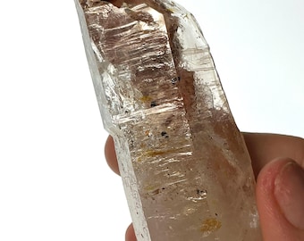 May include: A clear quartz crystal with a reddish-brown hue and inclusions. The crystal is pointed at the top and has a smooth, polished surface.