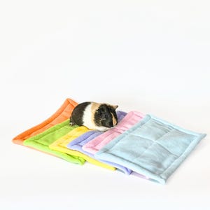 May include: A multi-colored guinea pig sits atop a stack of colorful, square pet bed pads. The pads are in shades of orange, green, yellow, purple, pink, and blue. The pads have a quilted design and are made of a soft material.