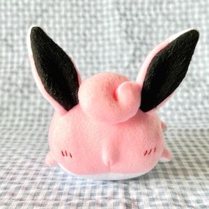 May include: A pink plush toy with large black and white ears. The toy has a round body, a curled detail on its head, and small red stitched details near its eyes. The toy is sitting on a checkered surface.