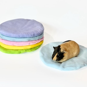 Puede incluir: Una pila de cinco camas redondas para mascotas de colores vivos hechas de tejido polar suave. Las camas son en tonos de morado, azul, rosa, amarillo y verde. Un pequeño conejillo de indias está acostado en una cama azul claro.