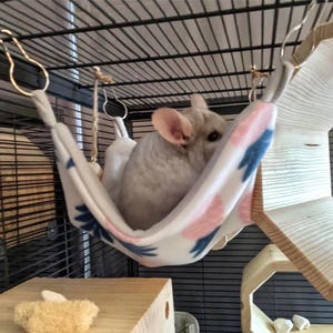 May include: A white and pink chinchilla hammock with blue floral designs hangs in a cage. A gray chinchilla is resting inside the hammock.