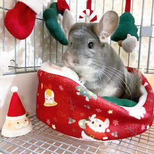 Könnte beinhalten: Ein graues Chinchilla sitzt in einem roten und weißen Weihnachts-Haustierbett. Das Bett hat eine weiße, flauschige Auskleidung und zeigt verschiedene festliche Designs, darunter Schneeflocken, Weihnachtsbäume und den Weihnachtsmann.