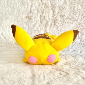 May include: A yellow Pikachu plush toy with brown ear tips and stripes on its back. The plush has pink cheek circles and is lying down on a white, fluffy surface. The toy is a popular character from the Pokémon franchise.