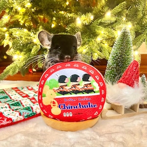 May include: A black chinchilla is holding a red and white round tin with the text "Butter Cookies Chinchulie" on it. The tin is decorated with a cartoon image of four soldiers in red uniforms and a Mickey Mouse-like character.