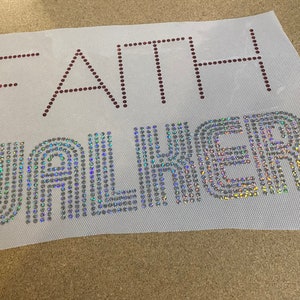 Könnte beinhalten: Ein weißer Stoff mit dem Namen "FAITH WALKER", der in Strasssteinen ausgeschrieben ist. Der Name "FAITH" ist in maronenfarbenen Strasssteinen, und der Name "WALKER" ist in schillernden Strasssteinen.