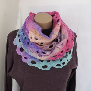 May include: A felted cowl scarf in shades of pink, purple, and blue. The scarf has a scalloped edge and decorative holes throughout. The cowl is displayed on a mannequin, showcasing its circular design and colour gradient.