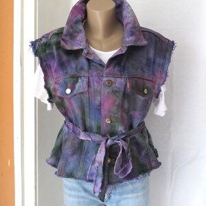 Può includere: Un gilet di jeans senza maniche con un motivo tie-dye nei toni del viola, blu e verde. Il gilet ha un colletto, chiusure con bottoni e una cintura abbinata. È abbinato a una camicia bianca e jeans azzurri.