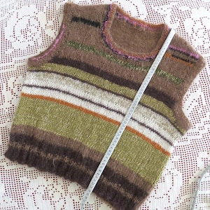 Puede incluir: Un chaleco sin mangas tejido a mano en tonos marrones, verdes y blancos con rayas horizontales. El chaleco tiene un cuello redondo y se mide con una cinta métrica. El chaleco mide aproximadamente 51 cm de largo.