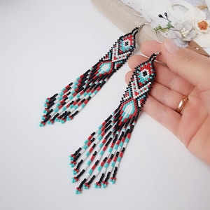 May include: Long, beaded earrings with a geometric pattern in black, white, red, and turquoise. The earrings are dangle style and have a fringe of beads at the bottom.