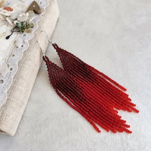 Pode incluir: Um par de brincos longos de contas vermelhas com efeito degradê. Os brincos são feitos com contas pequenas e redondas que são enfiadas juntas para criar um design tipo franja. As contas são de um vermelho intenso na parte superior e se desvanece gradualmente para um vermelho mais claro na parte inferior.