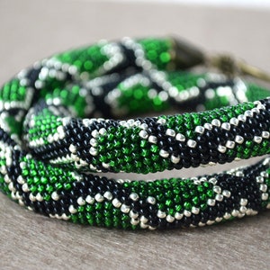 May include: A beaded bracelet with a striking pattern of green, black, and silver beads. The bracelet is coiled, showcasing the intricate design. The beads are small and tightly woven, creating a textured surface. The bracelet is a unique accessory.