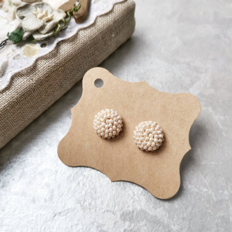 May include: A pair of round pearl earrings on a brown card. The earrings are made of small white pearls clustered together to form a round shape.