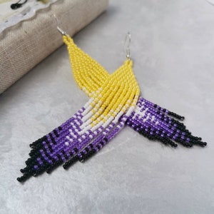 Può includere: Un paio di lunghi orecchini perlati in giallo, bianco e viola. Gli orecchini sono fatti di piccole perline rotonde e sono infilate insieme in un motivo a cascata. Gli orecchini sono una rappresentazione della bandiera del pride non binario.