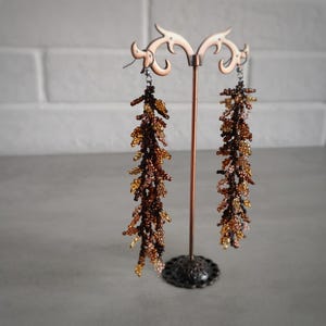 May include: A pair of long, beaded earrings in shades of brown and gold. The earrings are hanging from a metal stand.