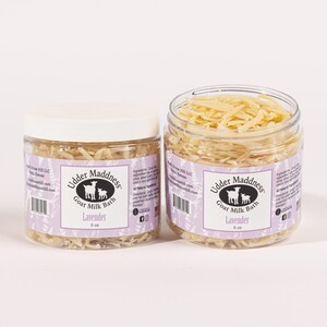 Peut inclure: Deux pots transparents de bain au lait de chèvre Udder Maddness, étiquetés "Lavender" et contenant 170 g chacun. Les pots sont remplis de matière séchée et déchiquetée. Les étiquettes sont lavande avec un logo blanc de chèvre et de chevreau.