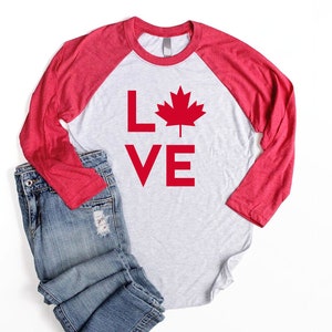 May include: A red and white long-sleeved baseball tee shirt with the words "LOVE" and a red maple leaf graphic. The shirt is laying on a white surface with a pair of blue jeans.
