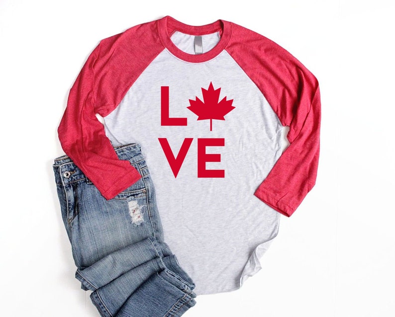 May include: A red and white long-sleeved baseball tee shirt with the words "LOVE" and a red maple leaf graphic. The shirt is laying on a white surface with a pair of blue jeans.