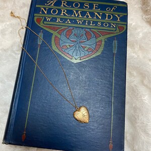 May include: A vintage book titled "A Rose of Normandy" by W.R.A. Wilson with a gold heart-shaped locket on a gold chain resting on top of it.
