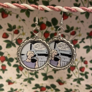 May include: A pair of silver-coloured dangle earrings. Each earring has a circular image with a comic strip illustration of a man and the text "WELL WHEN YOU SAY IT LIKE THAT IT SOUNDS STUPID." The earrings hang from silver hooks.