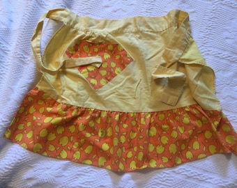 May include: A yellow apron with a ruffled bottom and a floral print pocket. The print features yellow flowers on a peach background.