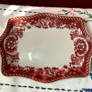 May include: A rectangular, white ceramic dish with a red floral design and a rope-like border. The floral pattern includes roses and other flowers. The dish is set on a white embroidered cloth.