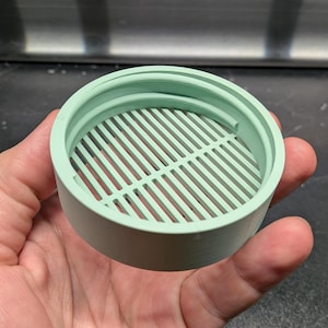 May include: A light green, cylindrical object with a slotted interior, held in a hand. The object appears to be made of plastic and has a circular shape with a series of parallel slots across its interior. The object is likely a filter or strainer.