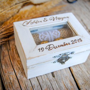 May include: A white wooden ring box with a glass top, engraved with "Ashton & Megan" and "19 December 2015." The box contains two gold rings and a lace detail. A silver clasp secures the box.