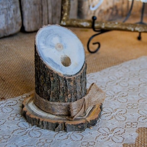 May include: A rustic wooden pen holder made from a section of a tree trunk, with a natural bark exterior and a hole for a pen. It sits on a wooden base and is adorned with a burlap ribbon. The background includes a sign that says "Our Guestbook".