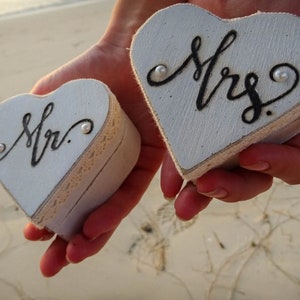 May include: Two white heart-shaped boxes with lids, one labeled "Mr." and the other "Mrs."  The boxes are made of wood and have a rustic finish. Each box has a small pearl embellishment.
