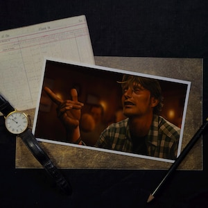 May include: A photograph of a man gesturing with his hand, wearing a plaid shirt. A gold wristwatch with a black leather band, a pencil, and a ledger are also visible. The image is set against a dark background.