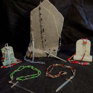 May include: An assortment of beaded necklaces with sword pendants. The necklaces feature various colors of beads, including black, blue, green, and red. The swords are silver-colored and are attached to the necklaces. The necklaces are displayed on a black surface.