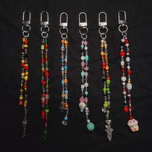 May include: Six beaded keychain charms with silver-tone clasps. Each charm features a unique combination of colorful beads, including red, yellow, green, and blue. Some charms have small decorative pendants, such as a knife, a fish, and a lucky cat.