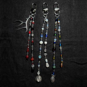 Może przedstawiać: Trzy breloki z koralikami i srebrnymi zapięciami wiszą na czarnym tle. Jeden brelok ma srebrny urok w kształcie poroża. Pozostałe dwa mają kolorowe koraliki w różnych kształtach i rozmiarach, w tym czerwone, niebieskie i przezroczyste.