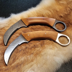 Op de afbeelding: Twee gebogen karambit-messen met lichtbruine houten handvatten en metalen vingerringen. Het bovenste mes heeft een donker, getextureerd lemmet met ingegraveerde kruisarceringen, terwijl het onderste mes een glad, gepolijst zilverkleurig lemmet heeft.