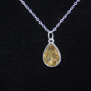 May include: A silver chain necklace with a teardrop-shaped pendant featuring a clear, amber-colored gemstone.