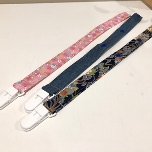 May include: Three fabric baby pacifier clips with white plastic clips. One clip has a pink floral pattern, one has a blue denim pattern, and one has a black and white floral pattern.