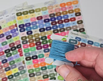 May include: A close-up of a blue embroidery floss wound on a white plastic bobbin. The bobbin has a label with the number "3810" printed on it. The bobbin is sitting on top of a chart of coloured embroidery floss.