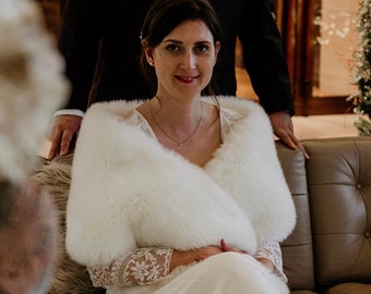 Puede incluir: Una novia con un vestido blanco y un chal de piel blanca se sienta en un sofá de cuero marrón. Un hombre con un traje negro se para detrás de ella. La pareja está en una habitación con un árbol de Navidad decorado con luces.