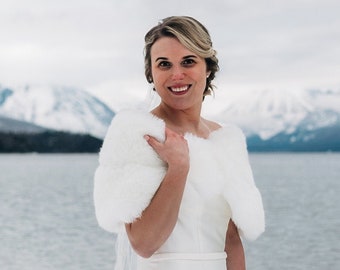 May include: A bride in a white wedding dress with a white fur shawl stands on a snowy beach with a bouquet of pink and purple flowers. The mountains and a lake are in the background.