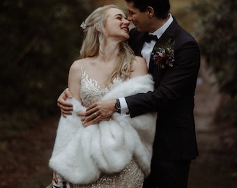 May include: A bride and groom are standing in a forest, looking at each other and smiling. The bride is wearing a white fur shawl over a beaded gown. The groom is wearing a black suit and a bow tie.