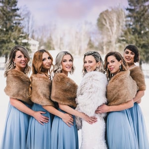 May include: Six bridesmaids in blue dresses with fur wraps stand in a snowy outdoor setting. The bride is in the center wearing a white dress with a fur wrap.