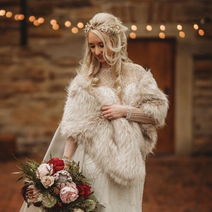 May include: A bride wearing a white wedding dress with a fur shawl and a bouquet of red, pink, and white flowers. She is standing in front of a stone building with string lights.
