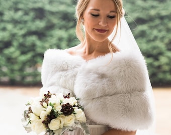 May include: A bride wearing a white wedding dress with a white fur shawl and a veil. She is holding a bouquet of white roses and greenery.